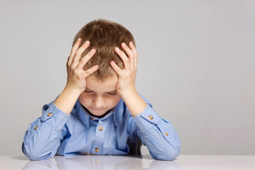 Upset young boy sitting at a table, holding his head with his hands